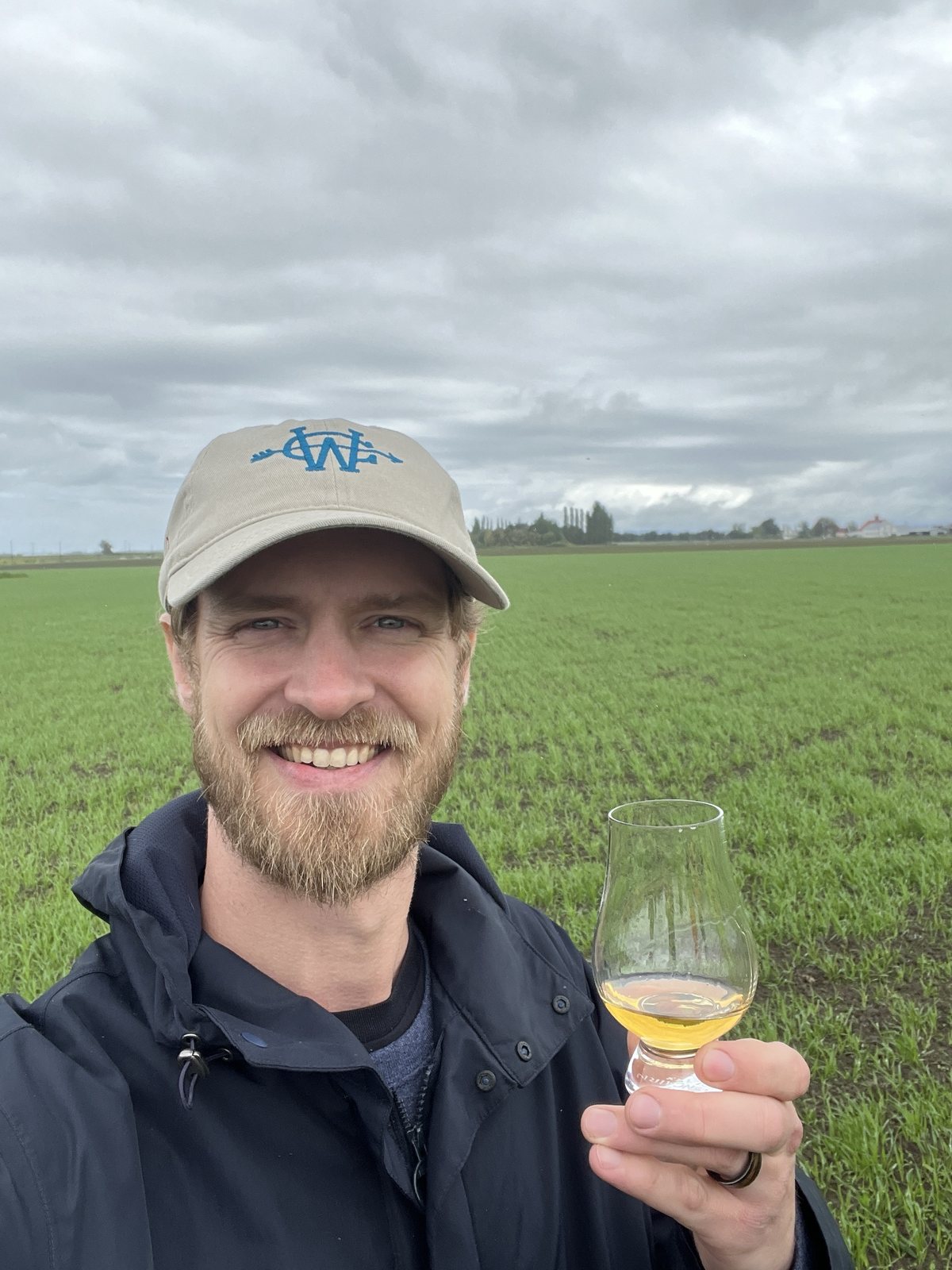 Tyler Pederson holding a glass of aged whiskey at the edge of the field of pilot-variety barley it was made from